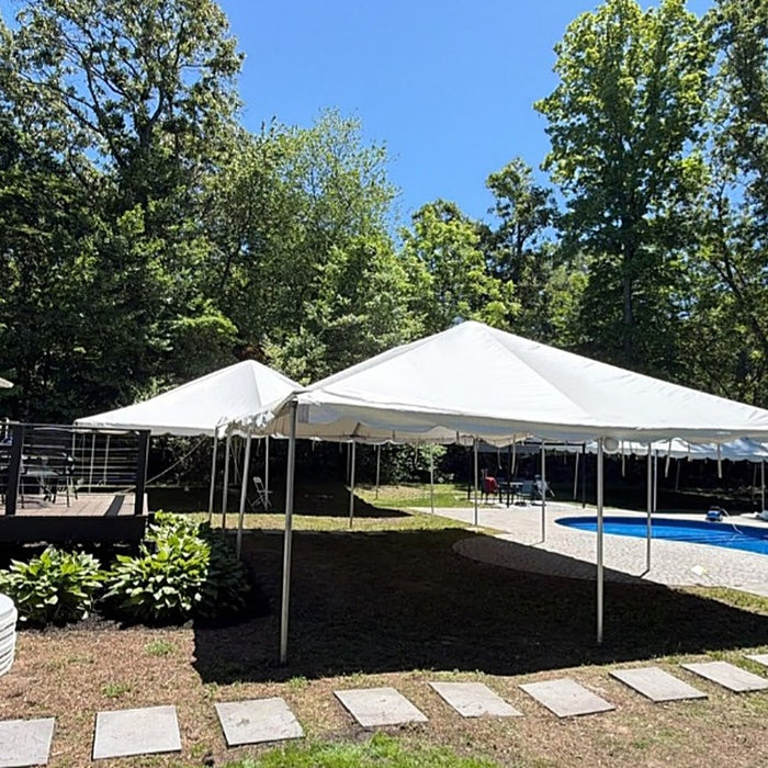 White frame party tents set up in a sunny backyard beside an in-ground pool and stone patio, with a deck, tall green trees and stacked round tables for event rental