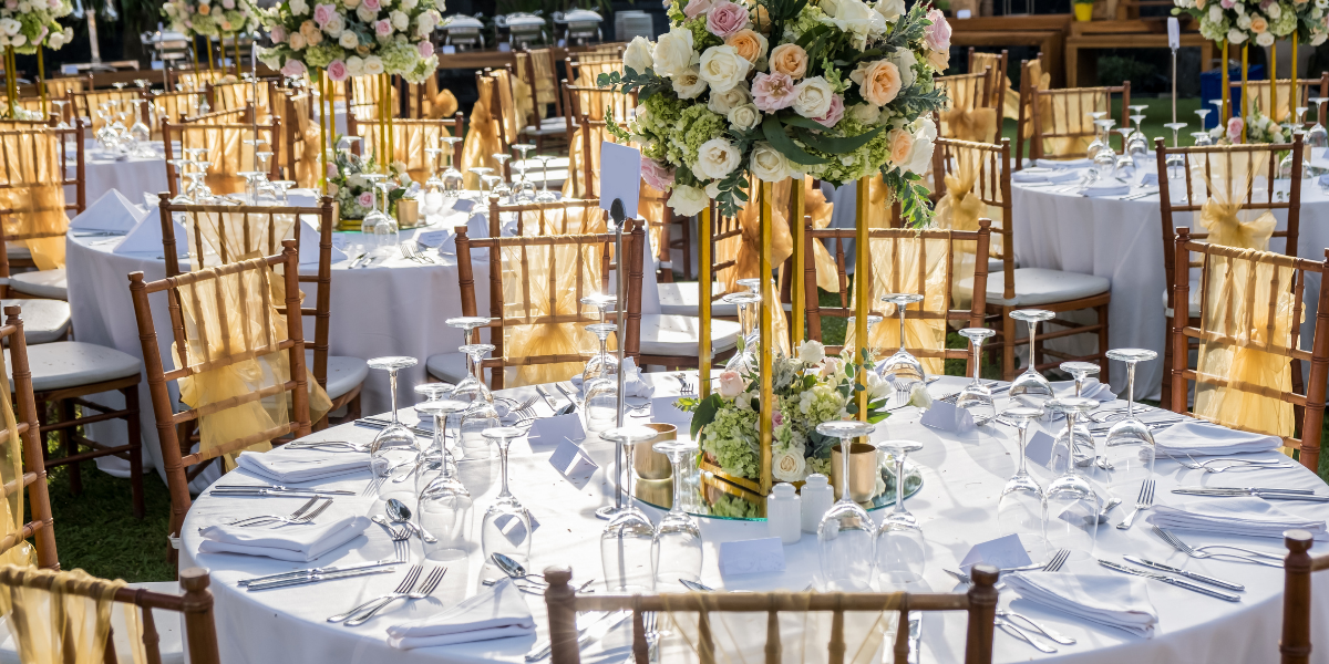 Round table set with white linens, gold-sashed chiavari chairs, and a tall floral centerpiece