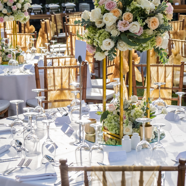Round table set with white linens, gold-sashed chiavari chairs, and a tall floral centerpiece