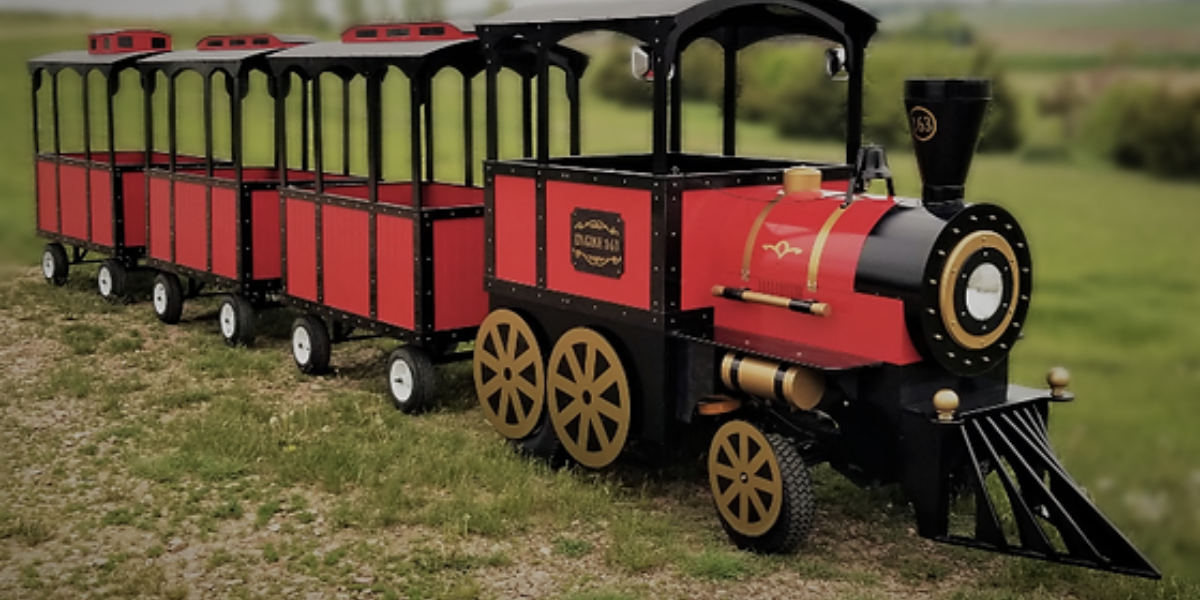 Red and black trackless tourist train with three open passenger cars parked on grass