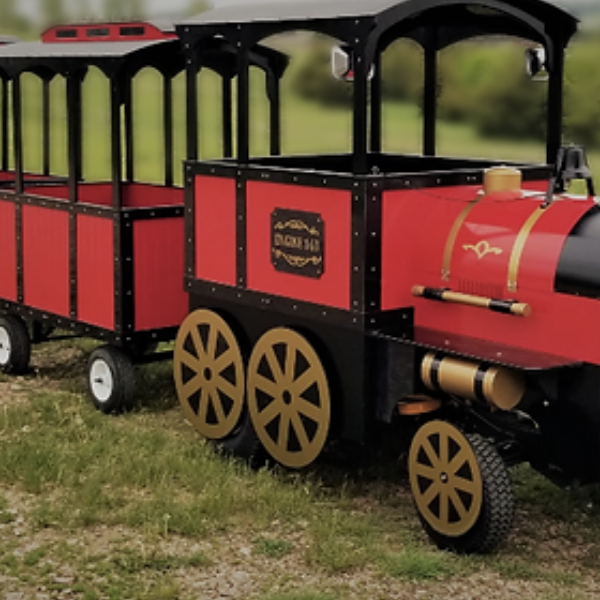 Red and black trackless tourist train with three open passenger cars parked on grass
