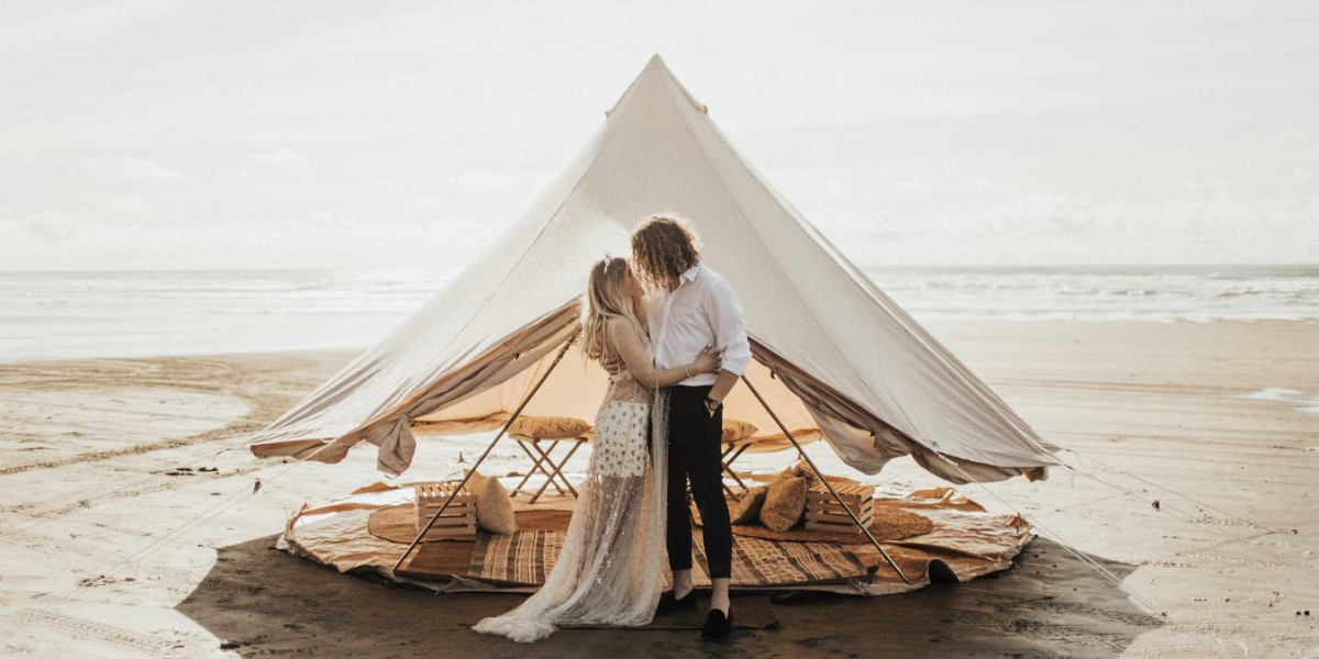 Cream bell tent on a beach with a couple embracing in front and the ocean behind them