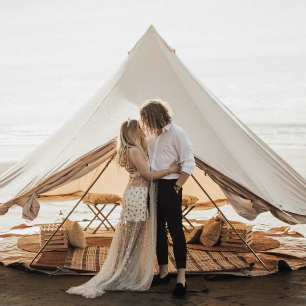Cream bell tent on a beach with a couple embracing in front and the ocean behind them