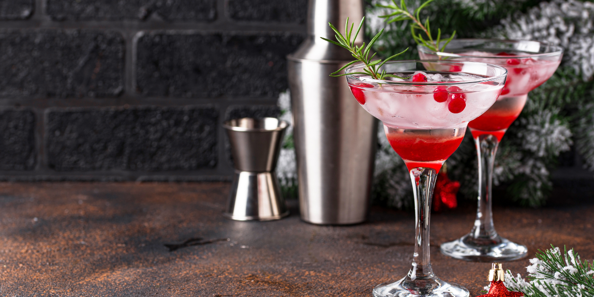 Two cranberry cocktails garnished with rosemary sprigs beside holiday pine decor