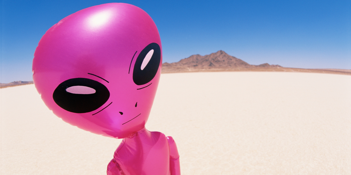 Pink inflatable alien figure standing on a flat desert salt pan with mountains behind