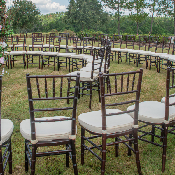 Rows of dark wood chiavari chairs with cream cushions curve around an outdoor ceremony