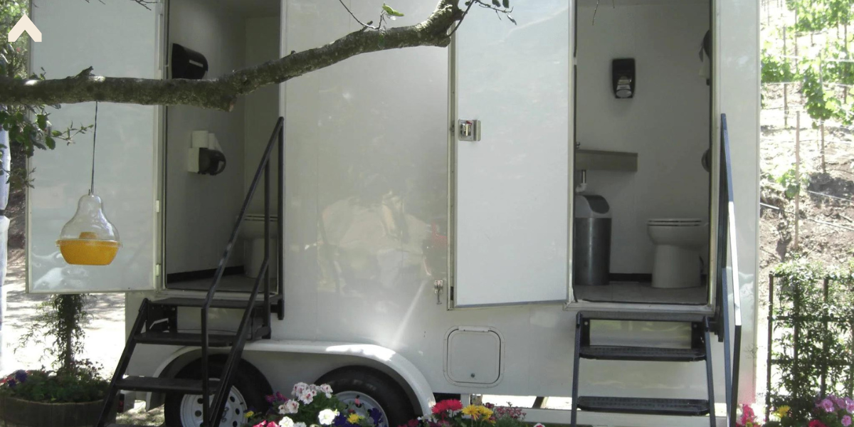 Portable restroom trailer with open doors showing toilets, flanked by flowers and a vineyard