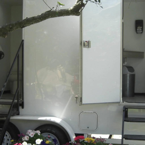 Portable restroom trailer with open doors showing toilets, flanked by flowers and a vineyard