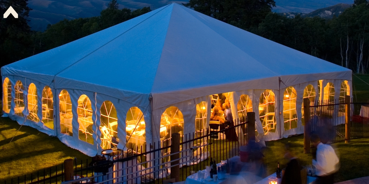 Large white pole tent glowing at dusk with cathedral windows and mountains behind it