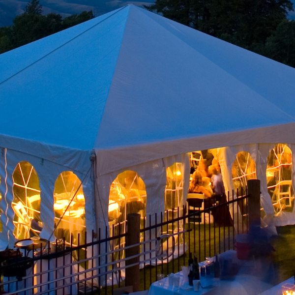 Large white pole tent glowing at dusk with cathedral windows and mountains behind it
