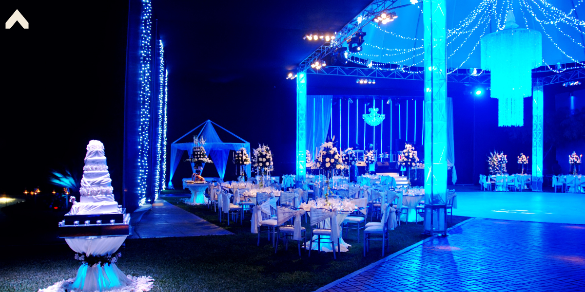 Blue-lit outdoor tent reception at night with round tables, chairs, and a tiered cake