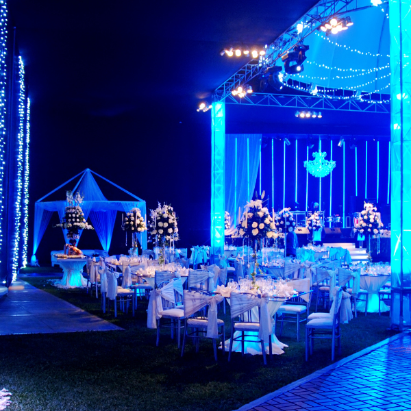 Blue-lit outdoor tent reception at night with round tables, chairs, and a tiered cake