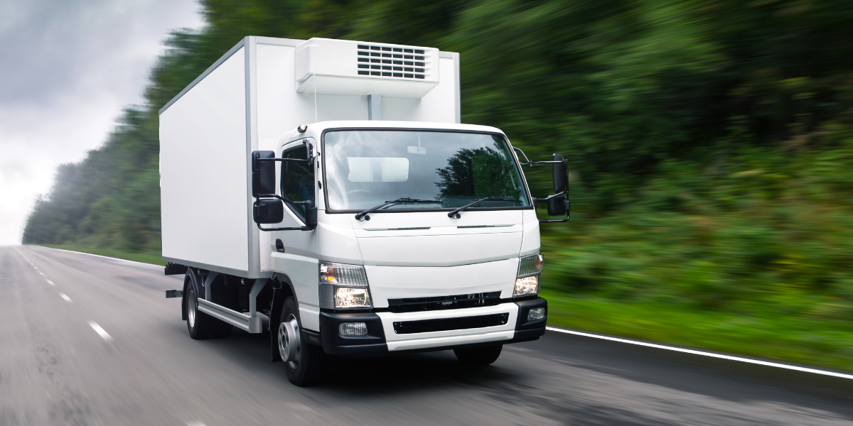 White box truck with a refrigerated cargo body driving along a forest-lined road