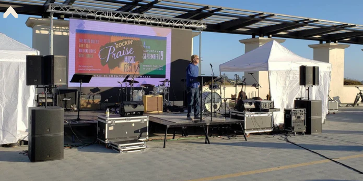 Musician on a raised stage platform with pop-up canopy tents nearby at an outdoor festival