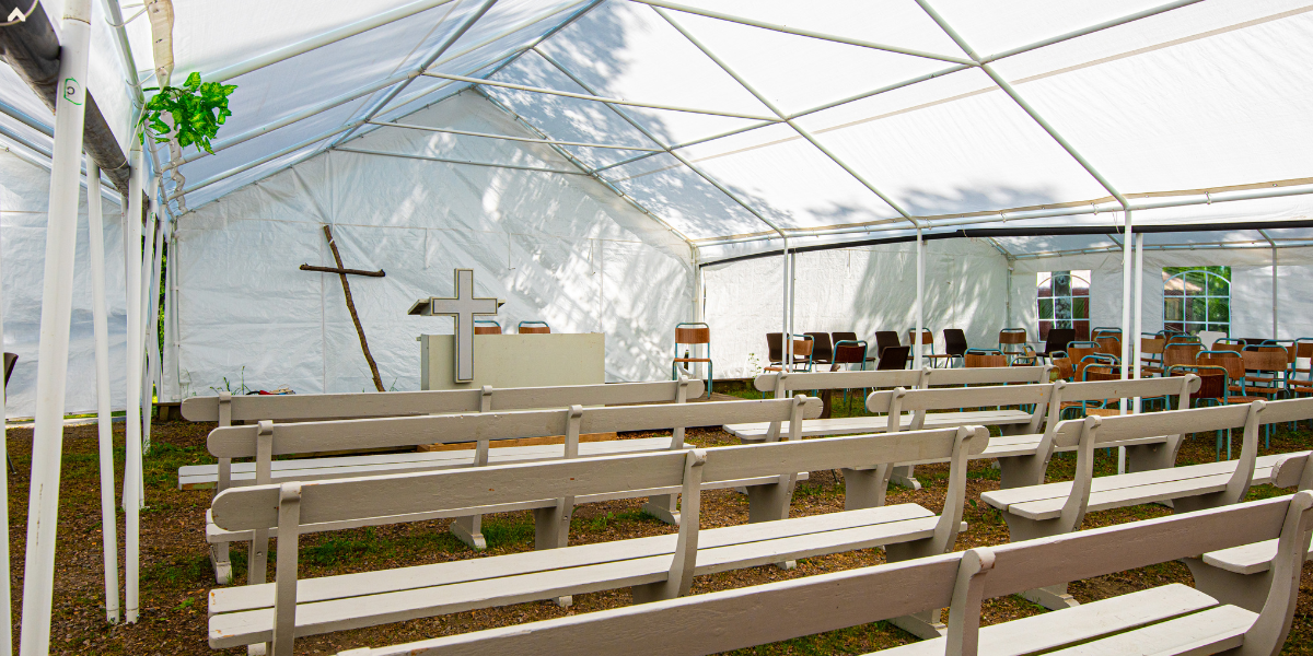 Interior of a white frame tent set up as an outdoor church with wooden pews and a cross