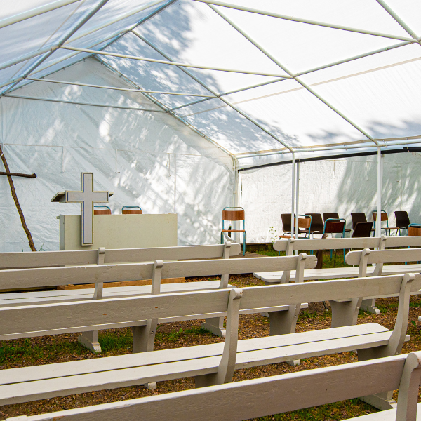 Interior of a white frame tent set up as an outdoor church with wooden pews and a cross