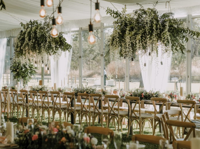 Tent interior with hanging greenery installations over a long table with cross-back chairs