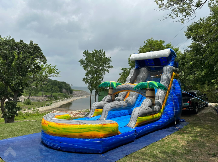 Tropical-themed inflatable water slide set up on a tarp overlooking a lake