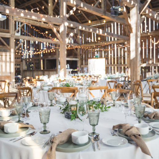 Rustic barn reception with round tables, cross-back chairs, and hanging string lights