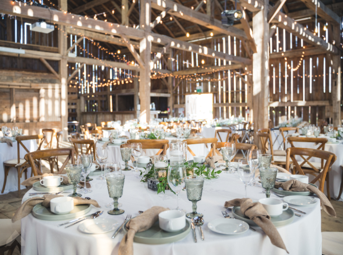Rustic barn reception with round tables, cross-back chairs, and hanging string lights