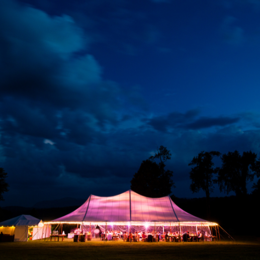 Illuminated peaked tent glowing at dusk with wedding reception tables set up inside