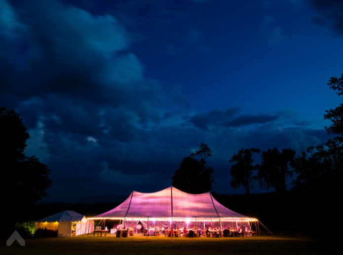 Illuminated peaked tent glowing at dusk with wedding reception tables set up inside