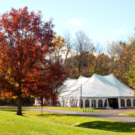 Large white pole tent with arched-window sidewalls set up amid colorful fall foliage