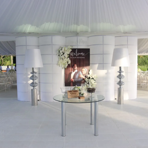 Tent entryway with glossy white flooring, draped fabric walls, and a round wedding welcome table