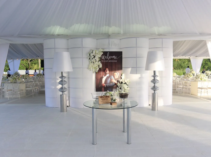 Tent entryway with glossy white flooring, draped fabric walls, and a round wedding welcome table