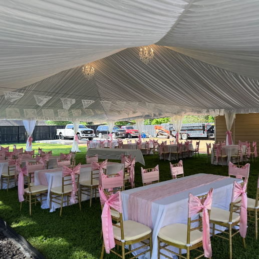 Tent interior with a draped fabric liner, mini chandeliers, and gold chairs with pink sashes