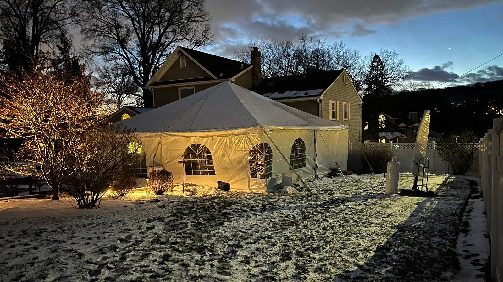 White frame event tent with arched window sidewalls lit from inside at dusk, set up in a snow-covered backyard beside a house for a winter event rental