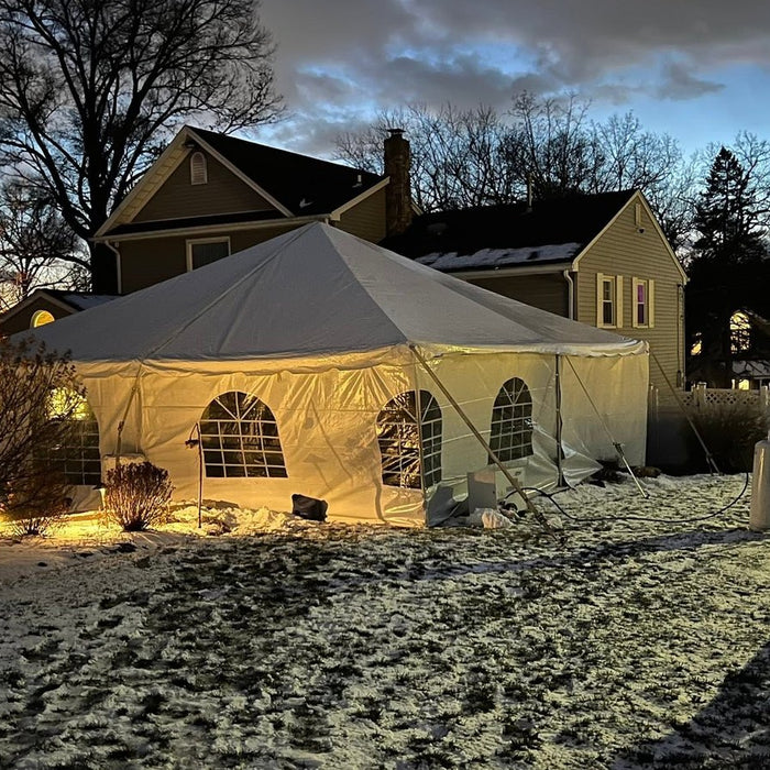 White frame event tent with arched window sidewalls lit from inside at dusk, set up in a snow-covered backyard beside a house for a winter event rental