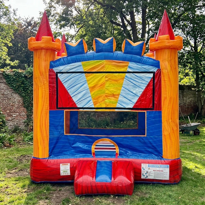 Red, orange, and blue castle-themed Royal Crown Bouncer inflatable set up on a grass lawn in a garden, with a brick wall and large tree in the background.