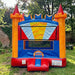 Red, orange, and blue castle-themed Royal Crown Bouncer inflatable set up on a grass lawn in a garden, with a brick wall and large tree in the background.
