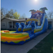 Tiki Tidal 17-foot commercial wet and dry inflatable water slide fully inflated in a residential backyard on a sunny day. Deep blue and purple tropical wave graphics, two green palm trees at the 17-foot peak, tiki climbing wall, single slide lane with water flowing, and a large rainbow-colored oval splash pool with yellow, orange, and red wave stripes at the base. Green grass and house with light siding in background.