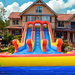 18' 2 Lane Unisex Wet & Dry Slide set up in a residential backyard, front view with ladder climb visible, large two-story house with balconies in background, blue sky with white clouds