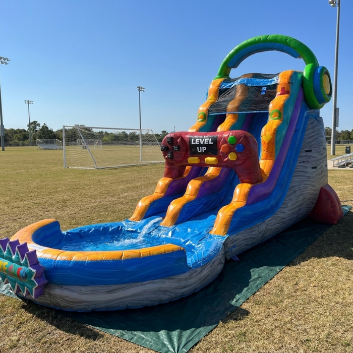 Real-world setup photo of the 18 foot Game Over wet and dry water slide filled with water and deployed on a grass sports field next to a soccer goal, shown under clear blue sky with bleachers visible in the background.