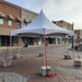 White frame event tent installed with concrete ballast weight blocks and red ratchet tie-down straps on a brick cobblestone street in a historic downtown district, with storefronts and awnings in the background.