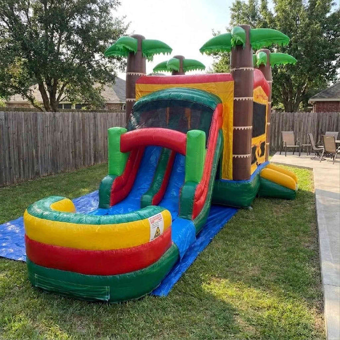 Tiki Trail Dual Lane Wet and Dry Combo set up on a blue tarp on green backyard grass. Full unit from front-left — dual water slide lanes and colorful splash pool on the left, large bounce house with yellow walls, mesh viewing window, and brown tiki column corners on the right, three green palm trees at the peak. Wooden privacy fence and patio chairs visible in background. Surrounding trees and overcast sky.