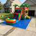 Tiki Trail Dual Lane Wet and Dry Combo fully inflated on a blue tarp in a residential concrete driveway. Front-left angle showing the complete 29-foot unit — dual water slide lanes and colorful splash pool on the left, large bounce house with mesh window and tiki-column corner trim on the right, three green palm trees on top. Beige house with attached garage and green lawn visible in the background. Blue sky.