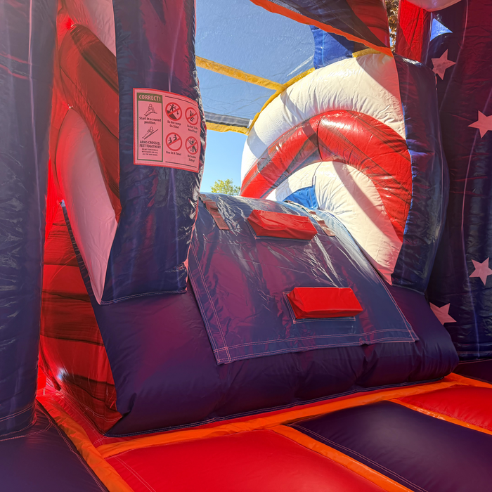Inside the American Titan Wet and Dry Combo: navy climbing ramp with red handholds, slide safety rules sign, red and navy bounce floor, white star walls and mesh roof netting