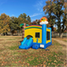 A professional installation of the Beyond Tent SolQuest 18oz Wet/Dry Bounce House Slide Combo unit in a park setting. It features vibrant blue, yellow, and red commercial-grade vinyl, an attached inflatable water slide leading into a splash pool, safety netting, and decorative elements like an inflatable palm tree and a smiling sun with sunglasses.