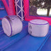 Close-up detail of the two large white round inflatable stabilizer cylinders at the base corners of the KPOP Combo Wet N Dry; pink and purple walls of the main bounce chamber visible in the background; green tree canopy partially visible above.