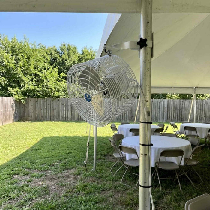 A large industrial floor fan with a white metal cage stands on grass next to a white event tent, with tables and chairs and a wooden fence in the background.