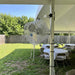 A large industrial floor fan with a white metal cage stands on grass next to a white event tent, with tables and chairs and a wooden fence in the background.