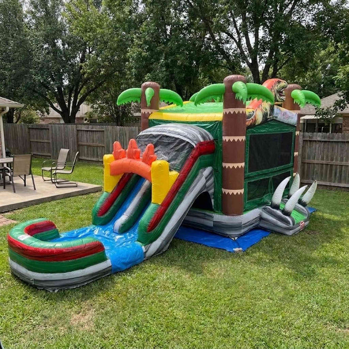 Beyond Tent Dino Stampede Bounce House Combo Wet & Dry set up in a residential backyard on grass, with a blue tarp under the slide landing area and a wood privacy fence in the background