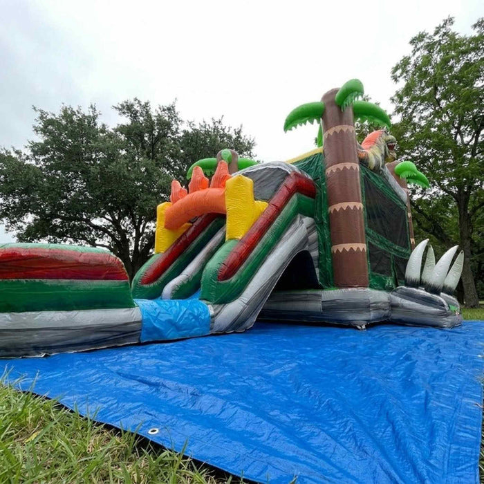 Beyond Tent Dino Stampede Bounce House Combo Wet & Dry, wide outdoor view on grass with a blue tarp beneath the unit and trees under an overcast sky