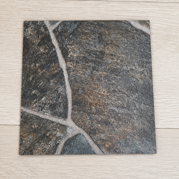 Close-up top view of a sample piece of Flagstone rolled vinyl flooring, featuring a dark gray and charcoal stone tile pattern with realistic light gray grout lines, displayed on a light oak wood grain background to show texture and thickness.