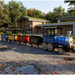 Frontier Trackless train blue and silver locomotive and four colorful passenger cars, parked on a cracked paved surface outdoors near a modern building with trees in the background.