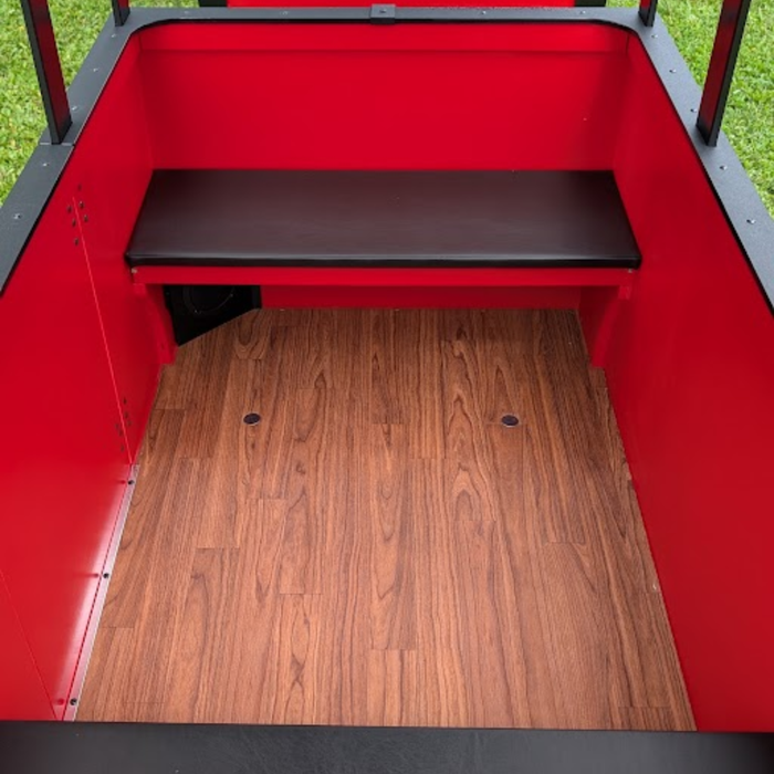 The back of a red trackless Frontier Train featuring a dark brown padded bench seat, a wood-look laminate floor, and red metal walls.